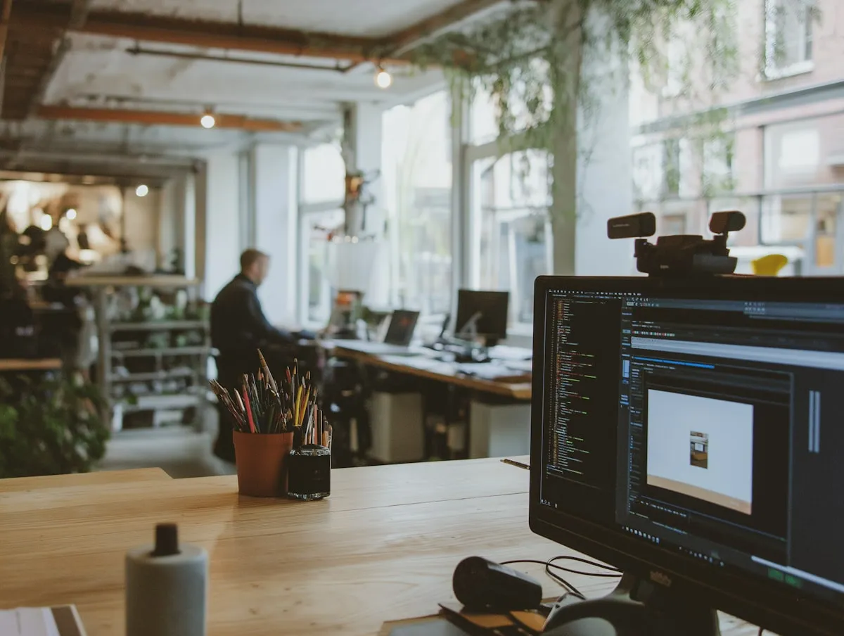 An open studio with a monitor on a wooden desk in the foreground and a colleague working by tall windows